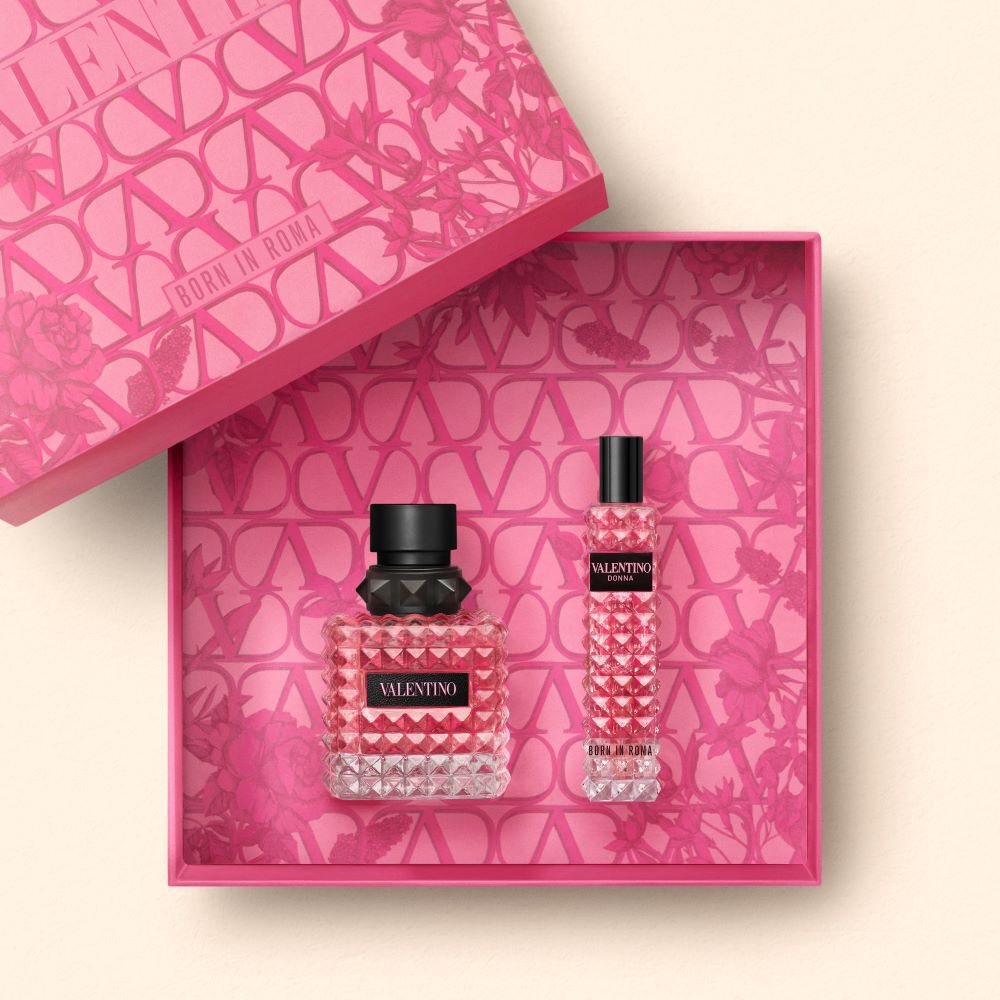 A stylish pink gift box featuring two Valentino perfume bottles: a larger one with a textured, crystal-like design and a smaller, matching bottle. The inside of the box is decorated with a vibrant floral pattern.