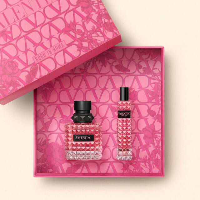 A stylish pink gift box featuring two Valentino perfume bottles: a larger one with a textured, crystal-like design and a smaller, matching bottle. The inside of the box is decorated with a vibrant floral pattern.