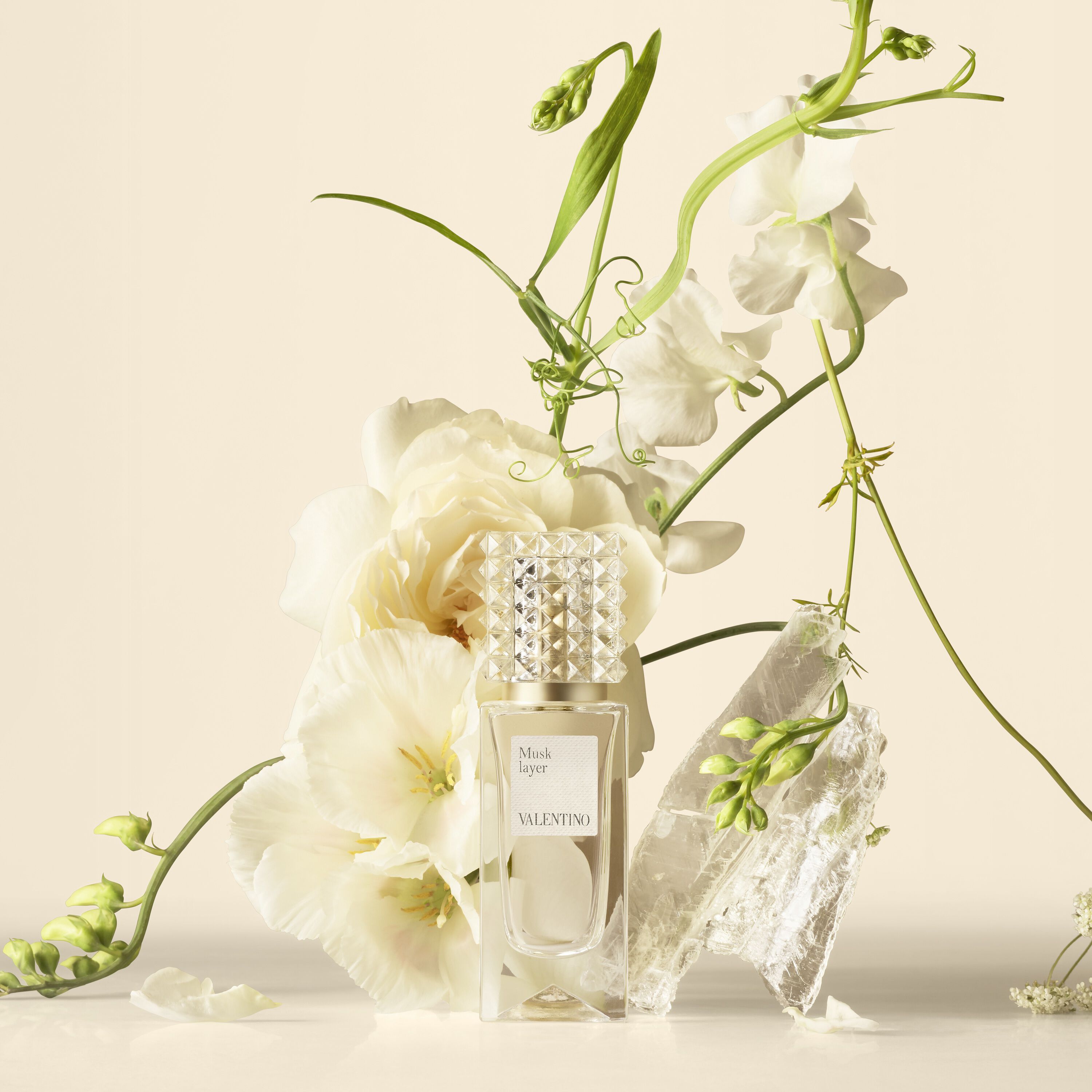 A Valentino perfume bottle arranged with white flowers and translucent crystals on a neutral background.