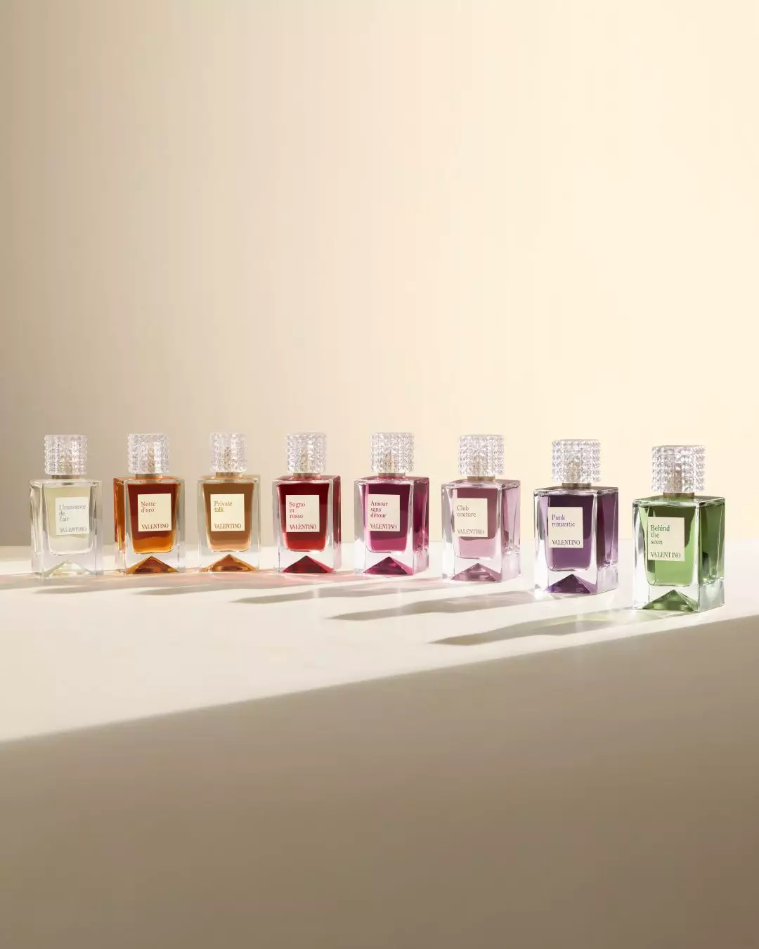 A row of elegant perfume bottles from Valentino, featuring various colorful scents, each with a textured lid and a label indicating its name, set against a soft, neutral background.