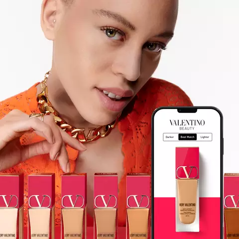 A model with short hair and a striking orange outfit poses next to a smartphone displaying Valentino Beauty foundation options, alongside several foundation bottles in varying shades.