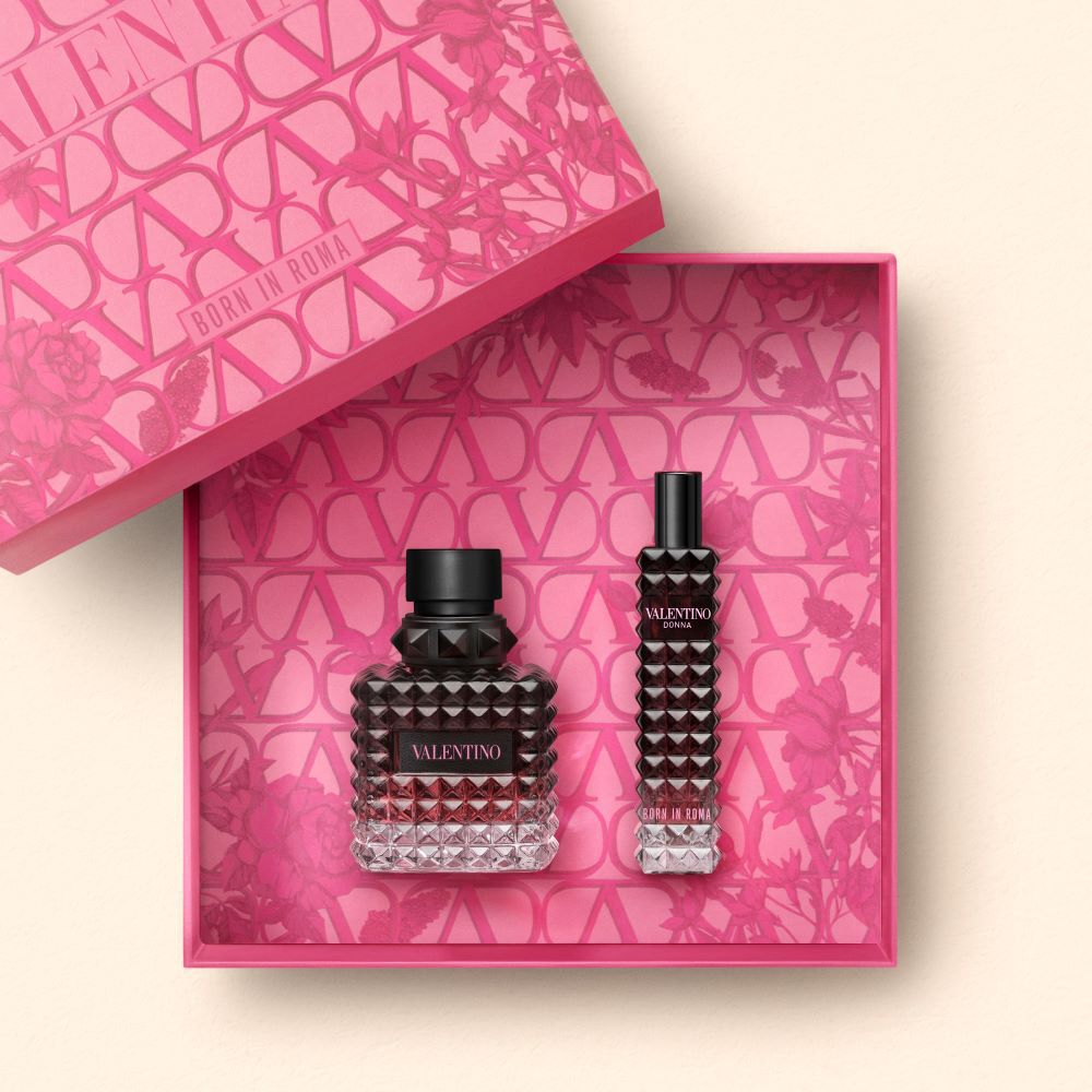 A beautiful presentation of Valentino perfume featuring a large, textured bottle and a smaller travel-sized vial, both displayed in a pink box adorned with floral and geometric patterns.