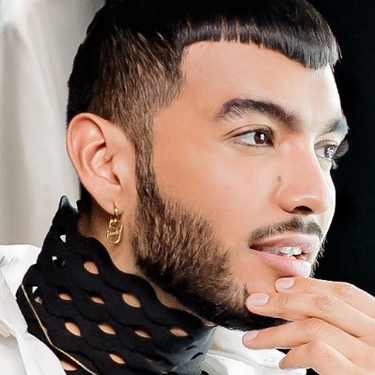 Un homme avec une barbe et une coiffure moderne, portant une chemise blanche et un foulard, se regarde dans un miroir avec un sourire pensif.
