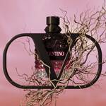 A stylish perfume bottle with a geometric shape, featuring a black cap and a pink label that reads "VALENTINO," is framed by abstract, twisted branches against a soft pink background.