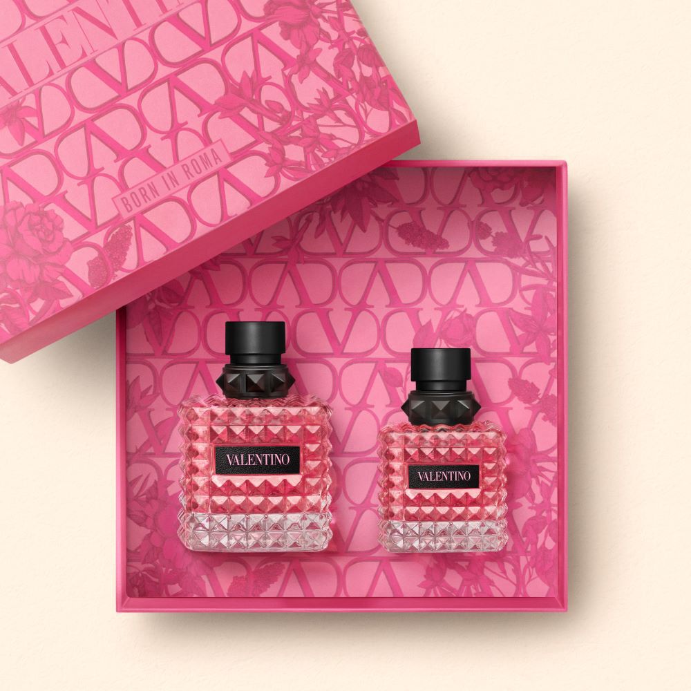 A pink gift box with a floral design contains two bottles of Valentino perfume, one larger and one smaller, both featuring faceted glass and black caps.