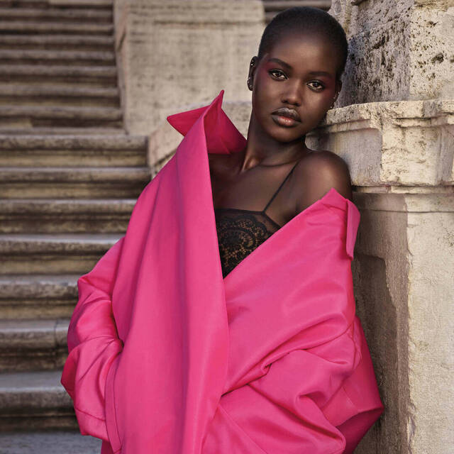 Une femme au teint fonce porte un manteau rose vif, posant avec elegance contre des escaliers en pierre.