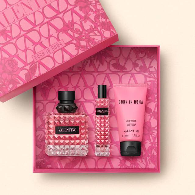 A pink gift box containing three Valentino beauty products: a large perfume bottle, a travel-sized perfume, and a scented lotion, all adorned with a diamond-patterned design.