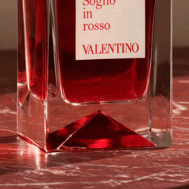 Un flacon de parfum Valentino, en verre transparent avec un liquide rouge a l'interieur, posant sur une surface en marbre.
