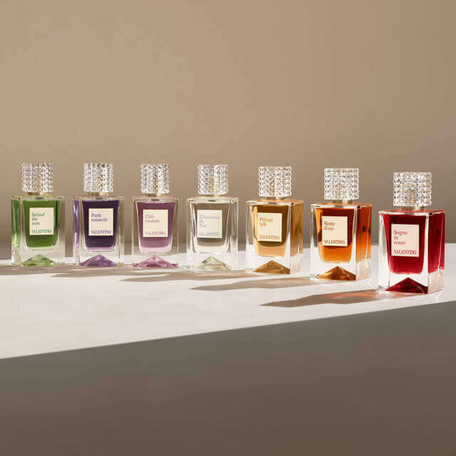 A row of elegant perfume bottles from Valentino, featuring various colorful scents, each with a textured lid and a label indicating its name, set against a soft, neutral background.