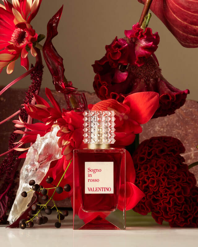 Un flacon de parfum Valentino Signo entoure de fleurs rouges eclatantes et de feuillage, sur un fond neutre.