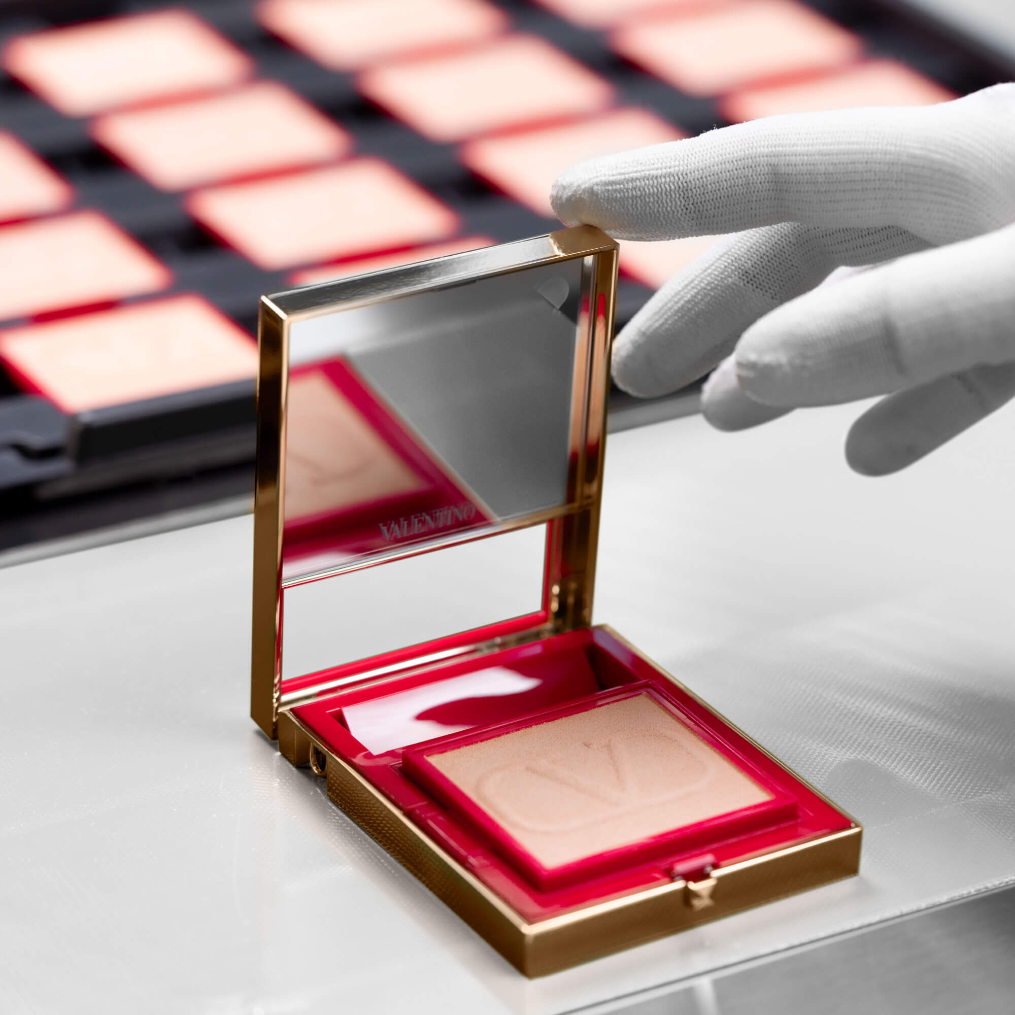A hand in a white glove is holding a gold compact mirror with a square blush inside, reflecting a clean, professional environment with blurred rows of product behind it.